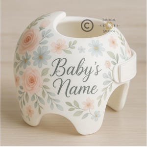 May include: A white baby helmet decorated with watercolor-style flowers in shades of pink and blue, with green leaves. The text "Baby's Name" is displayed in gray script. A white strap secures the helmet.