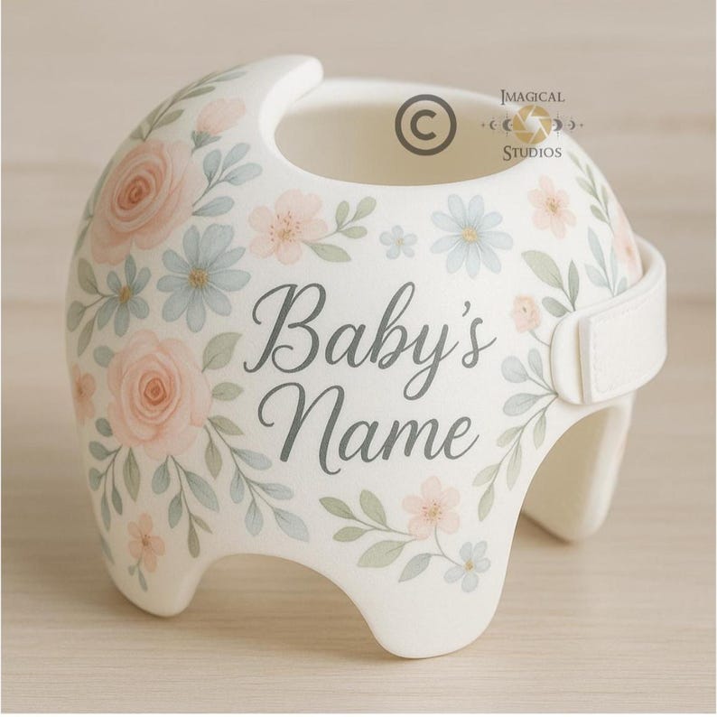 May include: A white baby helmet decorated with watercolor-style flowers in shades of pink and blue, with green leaves. The text "Baby's Name" is displayed in gray script. A white strap secures the helmet.