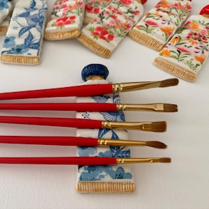 May include: A collection of ceramic paint tubes with floral designs in blue and red, and red-handled paint brushes with gold bristles. The brushes are arranged on top of a ceramic paint tube. The background is white.