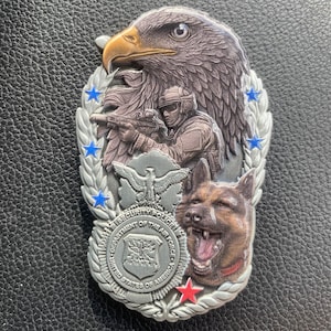 May include: A detailed silver-colored coin featuring an eagle, a soldier, and a German Shepherd. The coin has the text "SECURITY POLICE DEPARTMENT OF THE AIR FORCE UNITED STATES OF AMERICA" around the edge. Blue and red accents are visible.