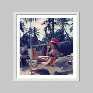 Pode incluir: Fotografia vintage de uma pessoa relaxando em uma espreguiçadeira, usando um chapéu vermelho e um traje de banho, segurando uma bebida. Palmeiras e outras pessoas são visíveis ao fundo, sugerindo um ambiente de resort tropical. Um balde de champanhe prateado está em primeiro plano.