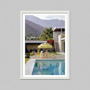 May include: A framed print of a vintage outdoor scene. A yellow umbrella shades a table where people are seated near a pool. A white dog stands nearby. Mountains and a modern house are in the background.