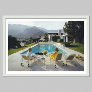 May include: A framed print depicting a mid-century modern scene. Two people are relaxing by a swimming pool, with lounge chairs and a small table. The scene is set against a backdrop of mountains and a clear blue sky. A woman in a yellow dress is sitting in a lounge chair.