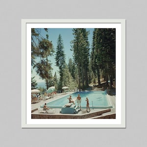 May include: A framed print of a vintage photograph featuring a rectangular swimming pool surrounded by people and tall pine trees. The pool is filled with clear water, and the sky is a bright blue. Lounge chairs and umbrellas are scattered around the pool.