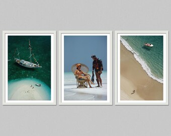 May include: Set of three framed art prints. One print shows a sailboat in turquoise water, another features a person in a wicker chair with a diver, and the third depicts a beach with a small boat and people.
