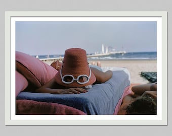 Peut inclure: Une personne allongée sur une serviette bleue à la plage, portant un chapeau bob en paille couleur terre cuite avec des lunettes de soleil à monture blanche posées sur le devant. La scène inclut un oreiller rose et une plage de sable.