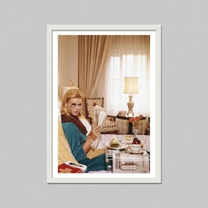 May include: A framed vintage photograph of a woman in a teal dress and brown fur collar, reading a newspaper with a monocle. A tray with food and drinks sits nearby. The room has a lamp, curtains, and a patterned chair.