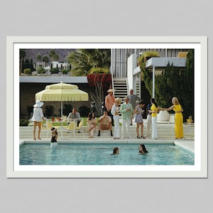 May include: A framed print of a vintage photograph depicting a poolside gathering. People in stylish attire are gathered around a swimming pool, with a yellow umbrella and palm trees in the background. The image evokes a retro, mid-century modern aesthetic.