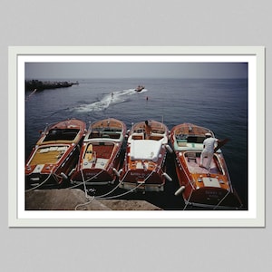 May include: Four classic wooden boats docked at a pier, with a speedboat in the distance. The boats are a rich mahogany color with white accents. A person in a white uniform stands on one of the boats. The water is a deep blue.