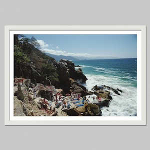May include: A framed photograph of a coastal scene with rocky cliffs, turquoise water, and a clear blue sky. People are gathered on the rocks, some near a shaded structure. The waves crash against the rocks, creating white foam.