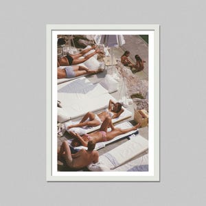 May include: A framed print featuring a sunny outdoor scene. Several people are sunbathing on white lounge cushions. Some are wearing swimwear. The image has a vintage aesthetic, with a focus on leisure and relaxation.