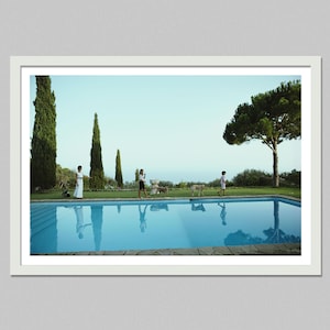 May include: A framed photograph of a swimming pool with clear blue water. People are walking on the grass near the pool, and their reflections are visible in the water. Tall trees and a clear sky complete the scene.