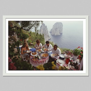 May include: A framed photograph of a group of people dining outdoors. The table is set with food and drinks, and the background features a scenic view of the ocean and cliffs. The people are dressed in casual attire.