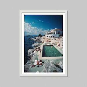 May include: A framed print of a coastal scene featuring a turquoise pool built into the rocky shoreline. A white building sits atop the cliffs, and people are seen sunbathing and swimming under a blue sky.