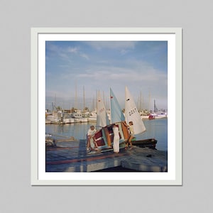 May include: A vintage photograph captures a sunny day at a marina. Several sailboats with white sails and wooden hulls are docked. People are seen handling a small sailboat on a wooden pier. The sky is blue with scattered clouds.