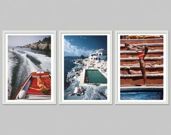 May include: Set of three framed art prints featuring vintage photographs. One shows a boat on the water, another a pool by the sea, and the third people sunbathing. The prints have white frames and a retro aesthetic.