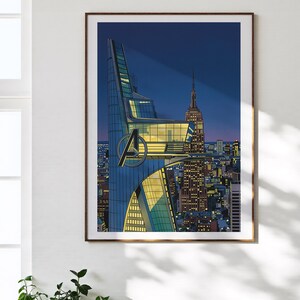 Op de afbeelding: Ingelijste kunstdruk met een gestileerde illustratie van een stadsgezicht 's nachts. Het kunstwerk toont een moderne wolkenkrabber met het Avengers-logo, naast het Empire State Building. Het kleurenpalet omvat blauw, geel en oranje.