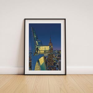 Op de afbeelding: Ingelijste kunstafdruk met een gestileerde stadsgezicht bij zonsondergang. De afbeelding toont een moderne wolkenkrabber met het Avengers-logo, het Empire State Building en een levendige skyline onder een diepblauwe hemel. Het kunstwerk zit in een zwarte lijst.