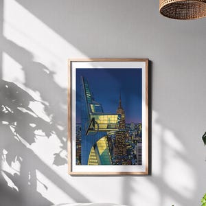 Op de afbeelding: Een ingelijste kunstdruk met een gestileerde stadsgezicht bij zonsondergang. Het kunstwerk toont een moderne wolkenkrabber met een onderscheidend ontwerp, tegen een achtergrond van een skyline met het Empire State Building. De print zit in een houten lijst.