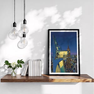 Op de afbeelding: Een ingelijste print met een stripachtige illustratie van een nachtelijk stadslandschap. Het kunstwerk toont een gebouw met het Avengers-logo, met de Empire State Building op de achtergrond. De print staat op een houten plank.