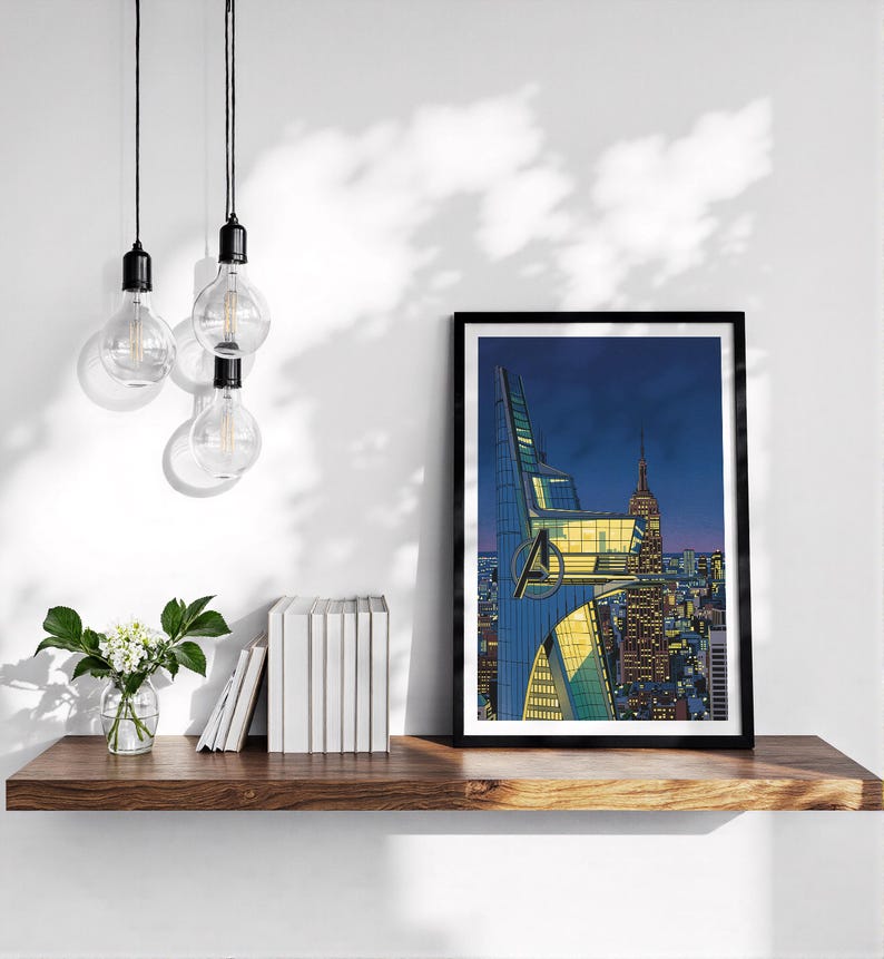 Op de afbeelding: Een ingelijste print met een stripachtige illustratie van een nachtelijk stadslandschap. Het kunstwerk toont een gebouw met het Avengers-logo, met de Empire State Building op de achtergrond. De print staat op een houten plank.