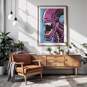 May include: A framed pop art print featuring a purple alien with a wide open mouth and sharp teeth against a light blue background. The artwork is displayed in a living room setting with a brown leather armchair and wooden cabinet.