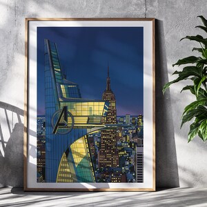 Op de afbeelding: Een ingelijste kunstdruk met een gestileerd stadsbeeld in de schemering. Het kunstwerk toont een moderne wolkenkrabber met het Avengers-logo, het Empire State Building en een levendige skyline onder een diepblauwe hemel. De print zit in een houten lijst.