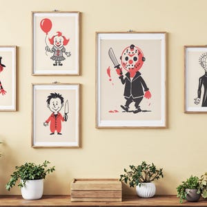 May include: A collection of framed art prints featuring cartoon illustrations of horror movie characters. The prints include a clown holding a balloon, a character with a hockey mask, and other figures in a vintage style. The art is displayed on a light yellow wall.