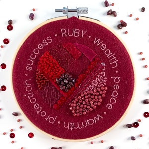 May include: A red embroidery hoop with a heart-shaped design in the center. The hoop is surrounded by a burgundy felt background with the words "Warmth", "Ruby", "Wealth", "Peace", "Protection", and "Success" embroidered around the edge.