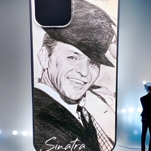 May include: A white phone case with a black and white illustration of Frank Sinatra wearing a hat. The case has a black border and the text "Sinatra" is written in white at the bottom. The text "Laura Label" is written in black at the top of the case.