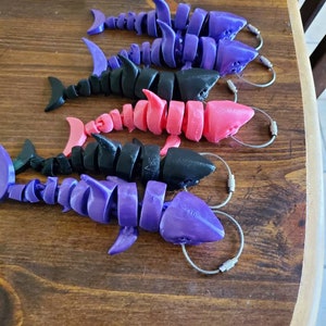 May include: Five articulated shark keychains in different colors. The keychains are made of plastic and have a silver metal ring. The sharks are in various shades of purple, black, and pink.