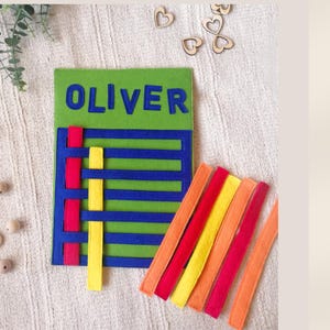Könnte beinhalten: Eine grüne Filztafel mit dem Namen "OLIVER" in blauen Buchstaben. Die Tafel hat blaue und rote gewebte Streifen mit gelben Streifen. Mehrere farbige Filzstreifen sind in der Nähe angeordnet. Holzperlen und Herzformen sind verstreut.