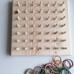 May include: A wooden pegboard with evenly spaced wooden pegs. A pile of colorful rubber bands in red, green, and tan is in the foreground. The pegboard is a light natural wood color.