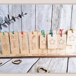 Puede incluir: Tabla de rutina de la hora de acostarse de madera con ocho etiquetas individuales. Las etiquetas están etiquetadas con actividades como "Usar el inodoro", "Baño", "Cepillarse los dientes", "Cepillarse el cabello", "Pijama", "Cuento para dormir", "Abrazo" y "Dormir". Cada etiqueta tiene un icono de estrella en la parte posterior.
