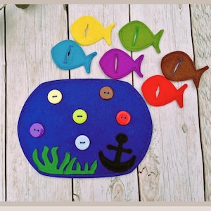 Könnte beinhalten: Ein Filzbrett mit einer blauen Ozeanszene, mit einem schwarzen Anker, grünen Algen und bunten Knöpfen. Es gibt sieben Filzfische mit Knopflöchern, jeder in einer anderen Farbe: gelb, blau, lila, grün, braun und rot.