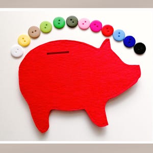 Puede incluir: Una alcancía de fieltro rojo con una ranura en la parte superior. La alcancía está rodeada de botones coloridos en forma de arcoíris.