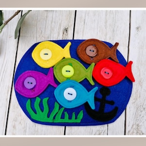 Puede incluir: Un tablero de fieltro con fondo azul que presenta seis peces coloridos con botones para los ojos. Los peces son amarillos, verdes, morados, azules, marrones y rojos. Hay algas verdes y un ancla negra en el tablero.