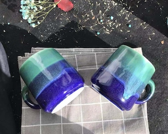 May include: Two ceramic mugs with a blue and green ombre glaze. The mugs have a rounded shape and a handle on the side.