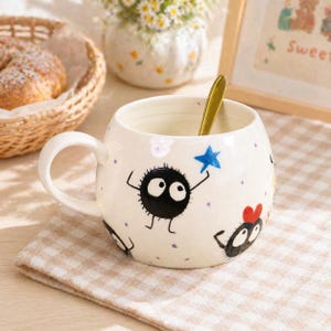 Puede incluir: Taza de cerámica blanca con un diseño caprichoso con sprites de hollín negros, una estrella azul y un corazón rojo. La taza tiene una cuchara dorada y un asa. La taza está sobre un paño a cuadros.