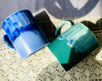 Op de afbeelding: Twee keramische mokken met een blauwe en groene glazuur. De mokken hebben een uniek, gemarmerd ontwerp met blauwe en groene strepen.