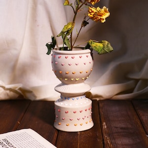 Pode incluir: Um vaso de cerâmica branco com um padrão de coração e uma base de pedestal. O vaso está cheio de rosas amarelas secas.
