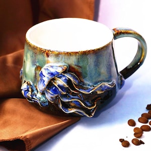 Puede incluir: Una taza de cerámica artesanal con un diseño único. La taza presenta un esmalte azul y verde con un diseño texturizado y esculpido en azul y dorado. Granos de café están dispersos cerca de la taza, que se encuentra sobre una tela marrón.