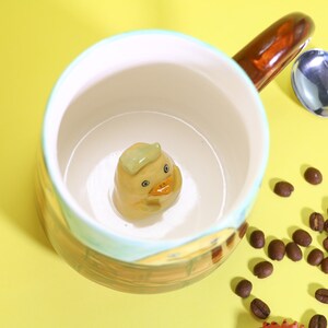 以下が含まれることがあります： 茶色の取っ手と、中に黄色いアヒルが入ったセラミック製のマグカップ。マグカップの外側には青と緑のデザインが施されています。コーヒー豆がマグカップの周りに散らばっています。