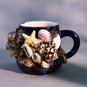 Puede incluir: Una taza de cerámica azul decorada con conchas marinas y una estrella de mar. La taza tiene un interior blanco y un asa azul.