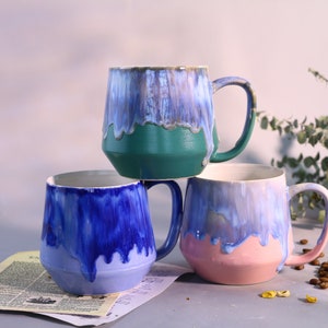 Puede incluir: Tres tazas de cerámica con efecto de glaseado de goteo. Las tazas están apiladas una encima de la otra. La taza superior es verde, la taza del medio es azul y la taza inferior es rosa.