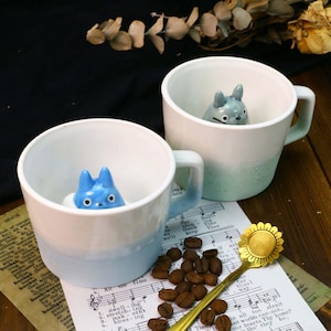 May include: Two ceramic mugs with blue and white ombre glaze. Each mug features a small ceramic figurine of a blue and white character inside. The mugs are sitting on a sheet of music with coffee beans scattered around them.