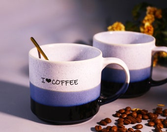May include: Two blue and white ceramic mugs with black rims. The mug on the left has the text "I ♥ COFFEE" printed on it. A gold spoon is inside the mug. Coffee beans are scattered on a grey surface.
