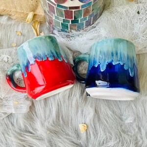 May include: Two ceramic mugs with a drip glaze design. One mug is red with a blue and green glaze, the other is blue with a green and white glaze.