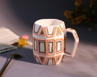 May include: White ceramic mug with a geometric pattern in orange, green, and pink. The mug has a unique, angular handle.