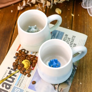 May include: Two ceramic mugs with blue and white glaze. Each mug features a small animal figurine inside. The mugs are resting on a newspaper with coffee beans scattered around them.